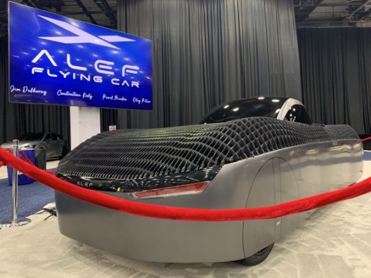 In the image above, we see the Alef Model A, a sleek and futuristic flying car, showcased at an event. The vehicle features an innovative design, notably lacking traditional wings, with a smooth, aerodynamic body. Its carbon fiber structure is evident in the grid-like pattern on the surface, emphasizing both its lightweight design and durability
