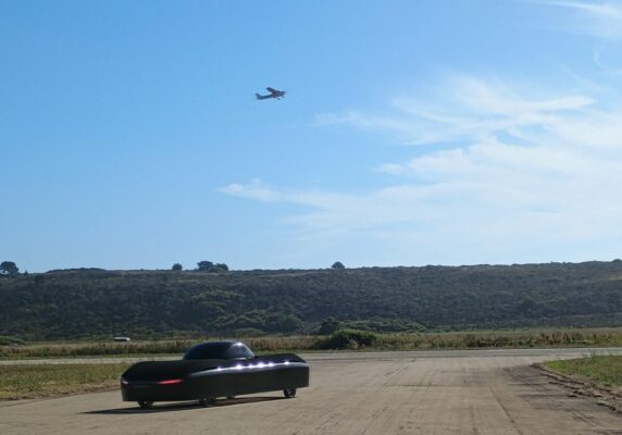The image depicts the flying car of Alef Aeronautics, a sleek, low-profile autonomous electric vehicle with a distinctive LED light array along its front, positioned on a rural runway